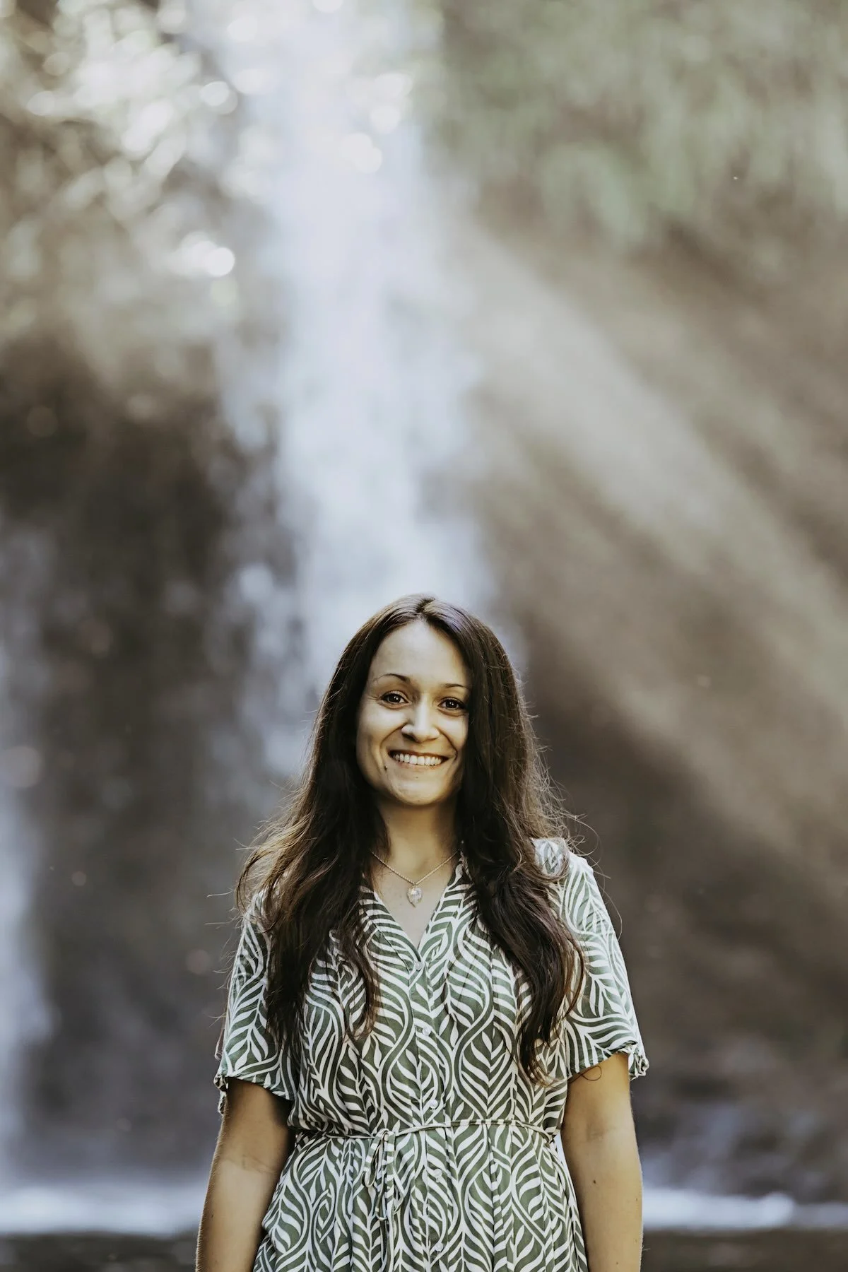 A smiling woman with long wavy brown hair wearing a green and white leaf patterned dress, standing outdoors in front of a blurred waterfall background.