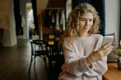 Young woman with curly hair smiling while looking at her phone in a cozy indoor setting, possibly a cafe or restaurant.