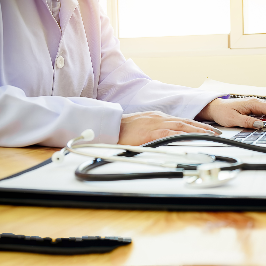 A healthcare professional wearing a white coat using a laptop at a desk, with a stethoscope and medical documents nearby.