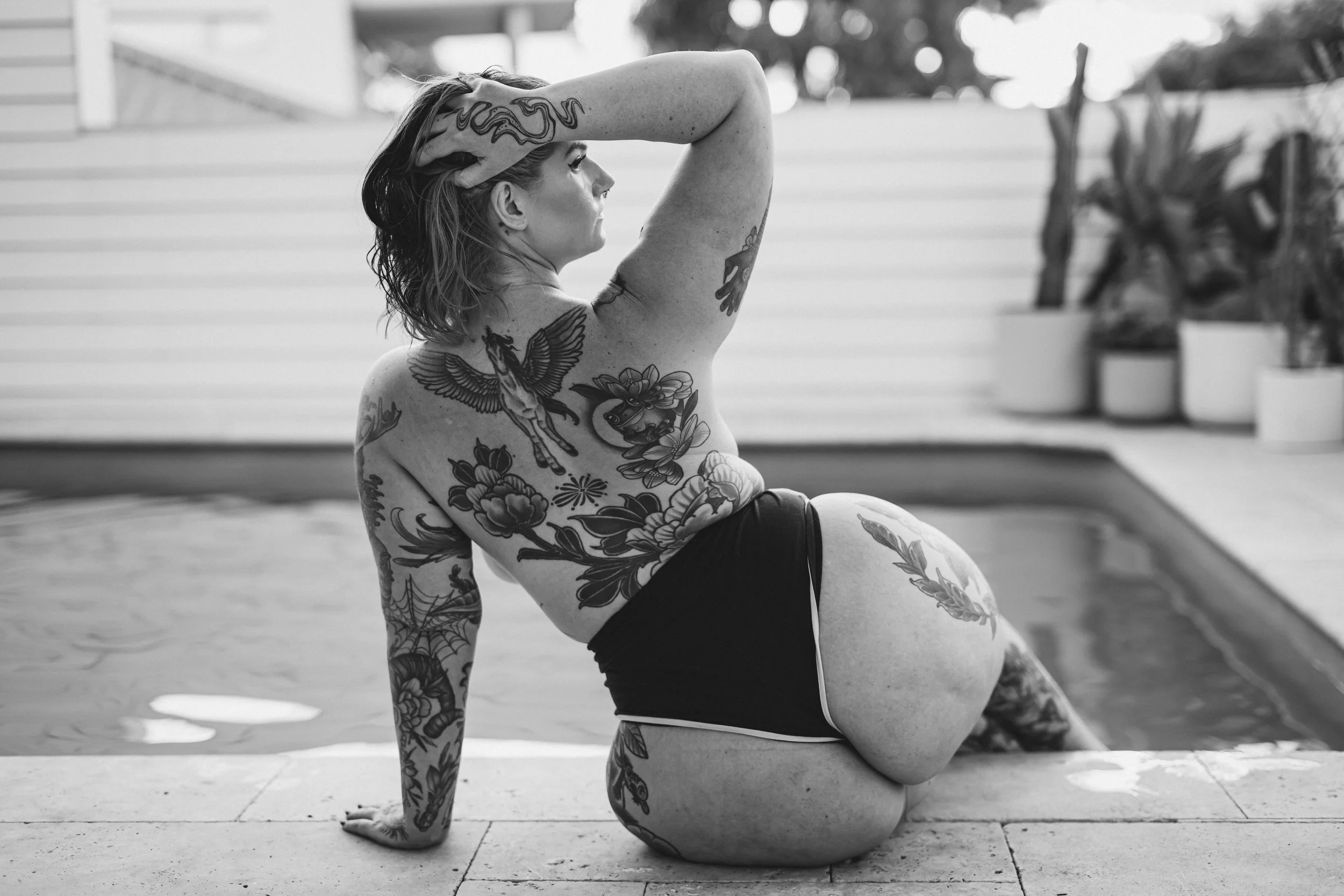 A woman with tattoos performing a yoga pose outdoors on a paved surface, with potted plants and a porch in the background.