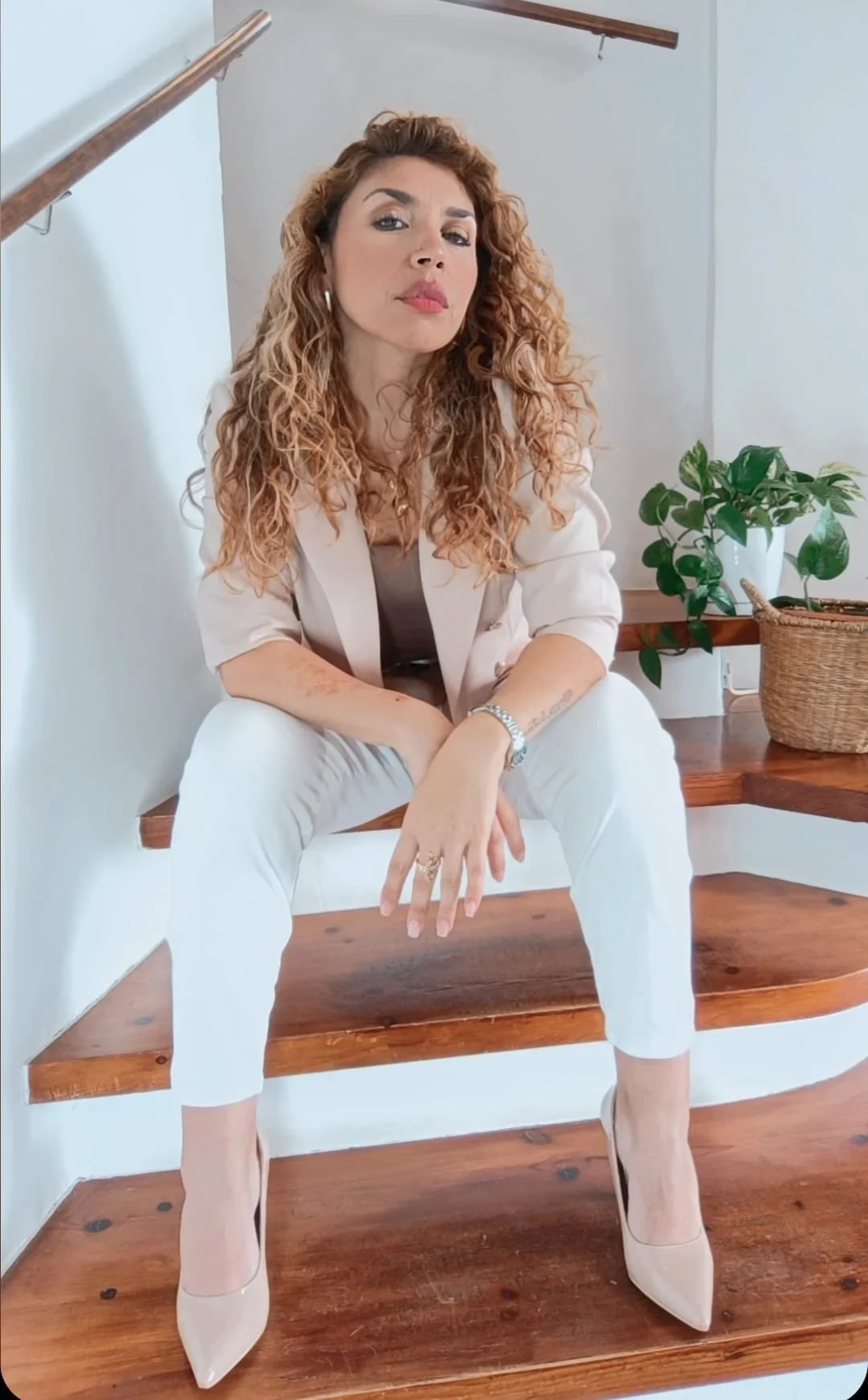 Mujer de cabello rizado y rubio, sentada en una escalera de madera, usando ropa beige y pantalones blancos, con plantas y pared blanca de fondo.
