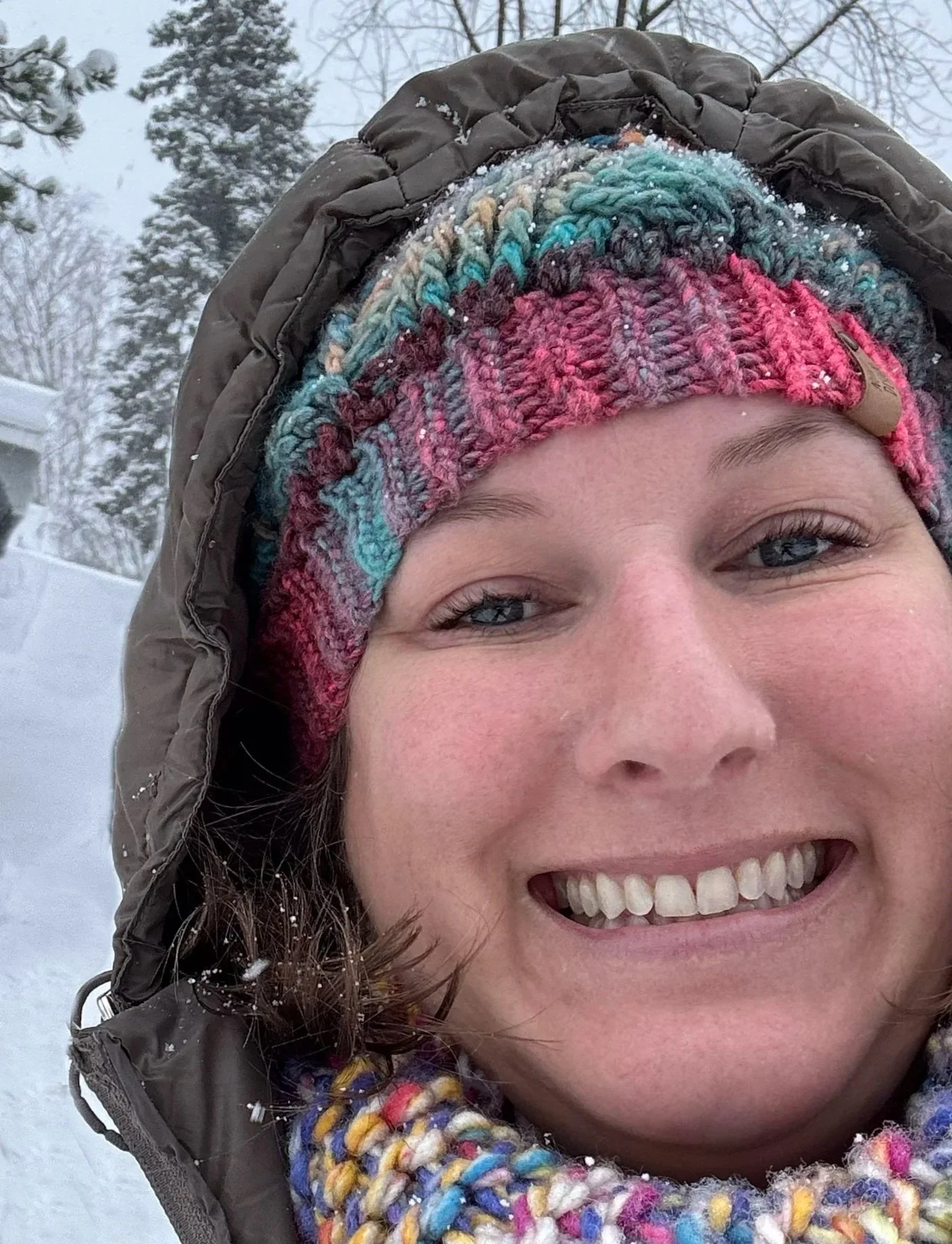 Close-up selfie of a smiling woman wearing a multicolored knitted hat, winter coat, and scarf in a snowy outdoor setting.