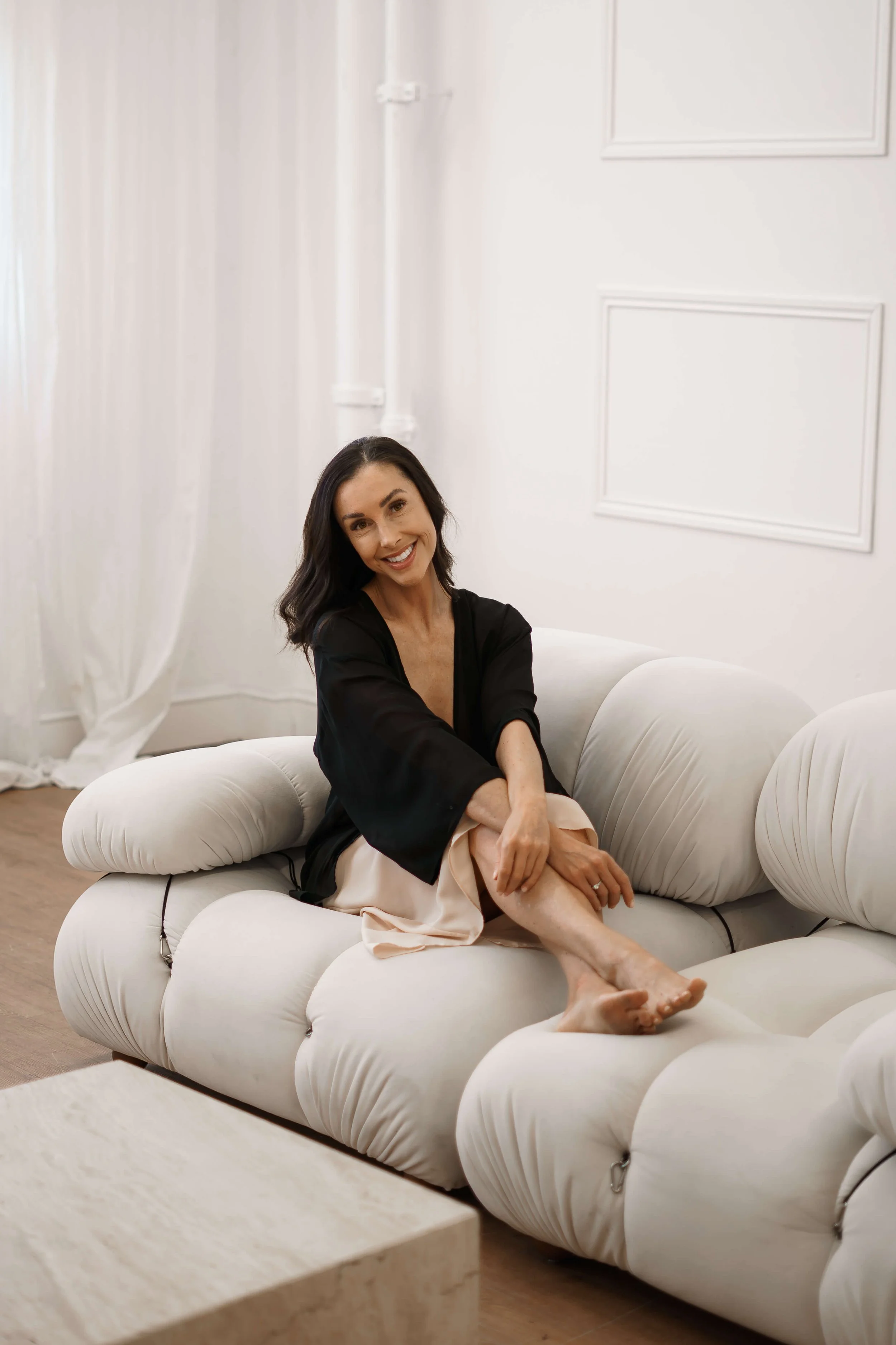 Smiling woman sitting on a plush white sofa in a minimalist room.