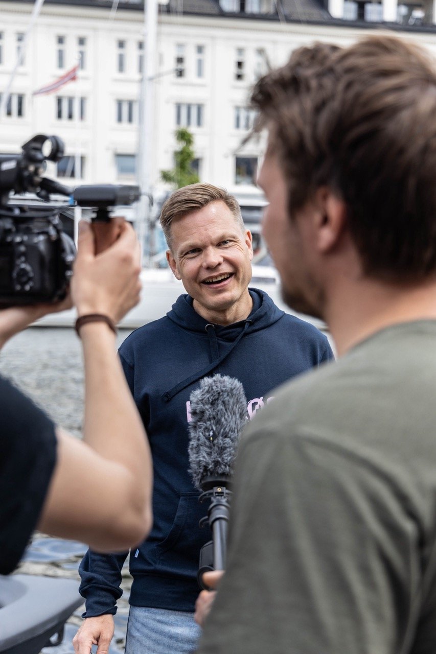 En mann som snakker med pressen, omgitt av to journalister med mikrofoner og kamera, utenfor en bygning.