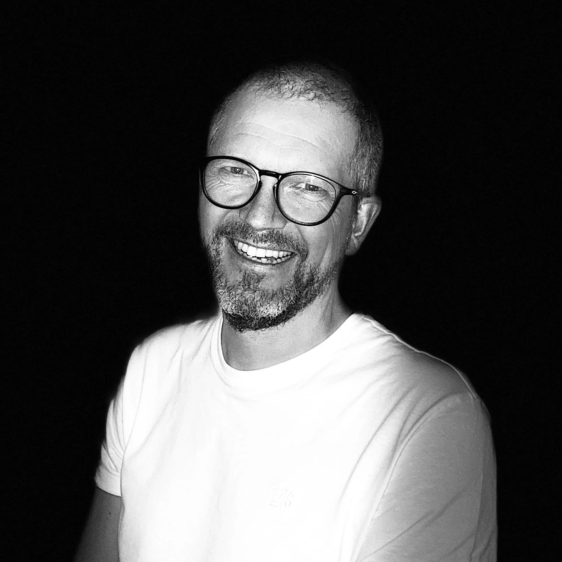 Svart-hvitt bilde av en smilende mann med kort hår, skjegg og glasses, iført hvit t-skjorte, mot en svart bakgrunn.