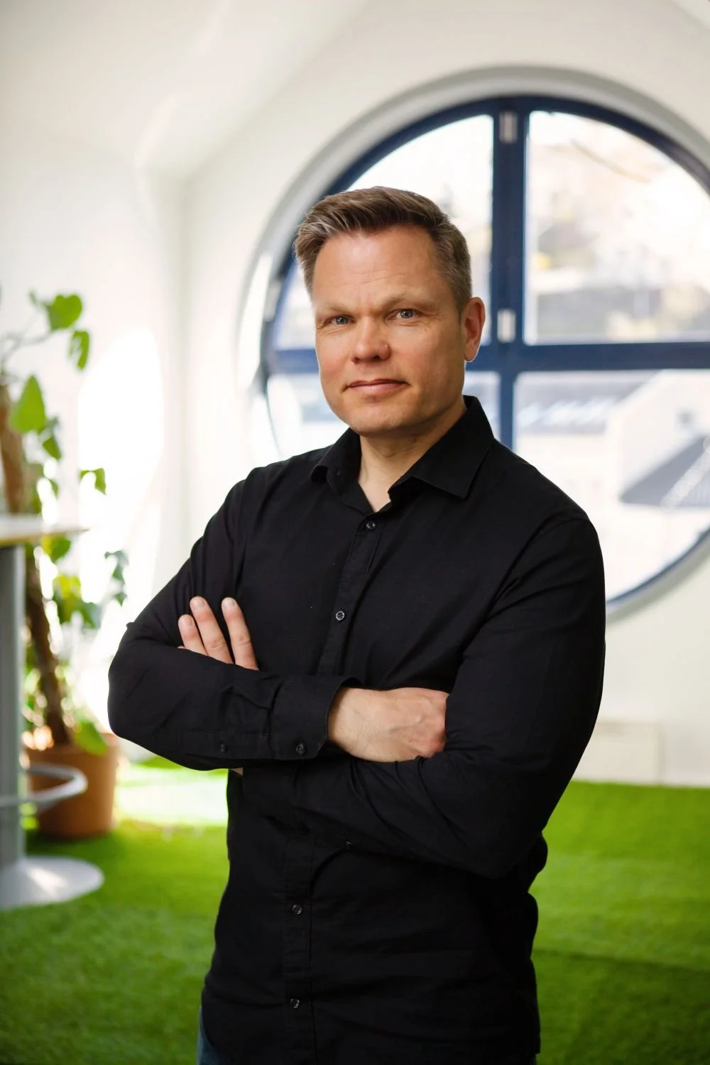 En mann i svart skjorte står med armene i kors foran et stort, rundt vindu i et lyst rom med grønne planter og kunstige gressmatter.
