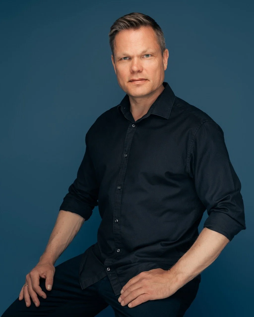 Portrett av en mann med kort, lys hår, i mørk skjorte, sittende mot en blå bakgrunn.