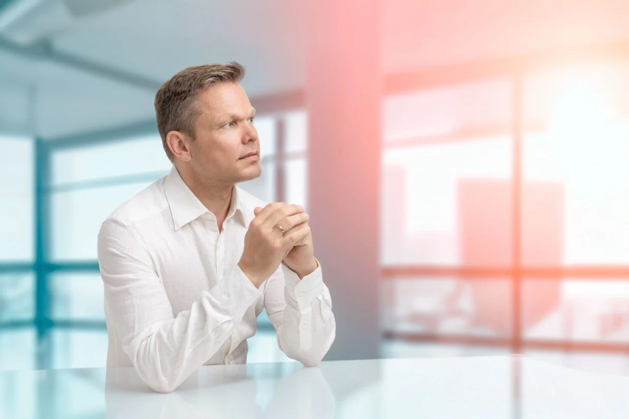 En man i hvit skjorte som sitter ved et bord i et moderne kontorlokale, ser ut til å tenke eller be om noe, med en lys og åpen bakgrunn.