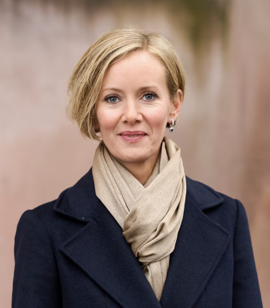 En kvinne med kort blond hår, iført en mørk kåpe og en lys fuskeskjerf, står foran en utendørs bakgrunn.