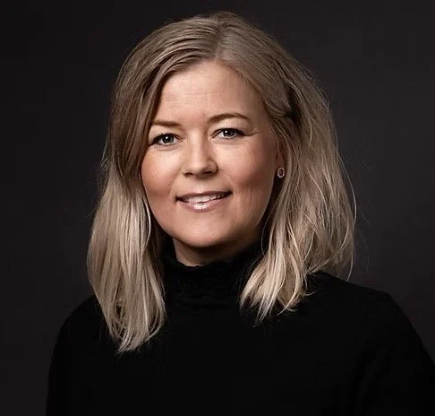 Portrett av en kvinne med skulderlangt blondt hår iført svart genser mot en svart bakgrunn.