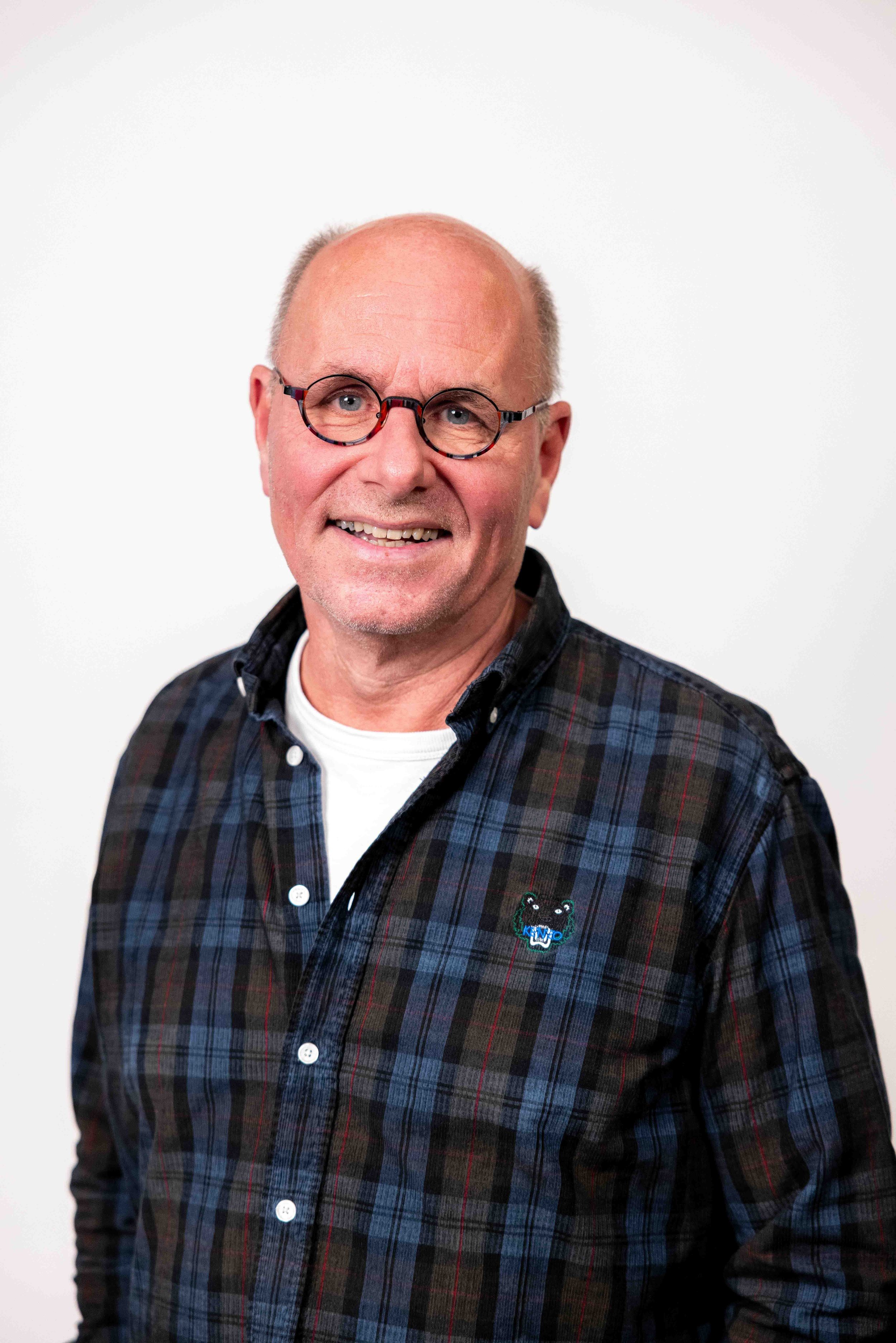 Et bilde av en smilende eldre mann med briller, iført en mørk rutete skjorte med en liten bjørneapplikasjon på brystet, foran en hvit bakgrunn.