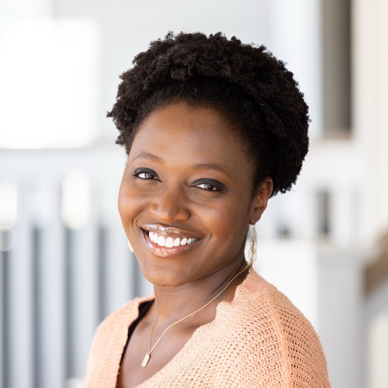 Et portrett av en smilende svart kvinne med kort, krøllete hår, iført en lys rosa genser og smykker.