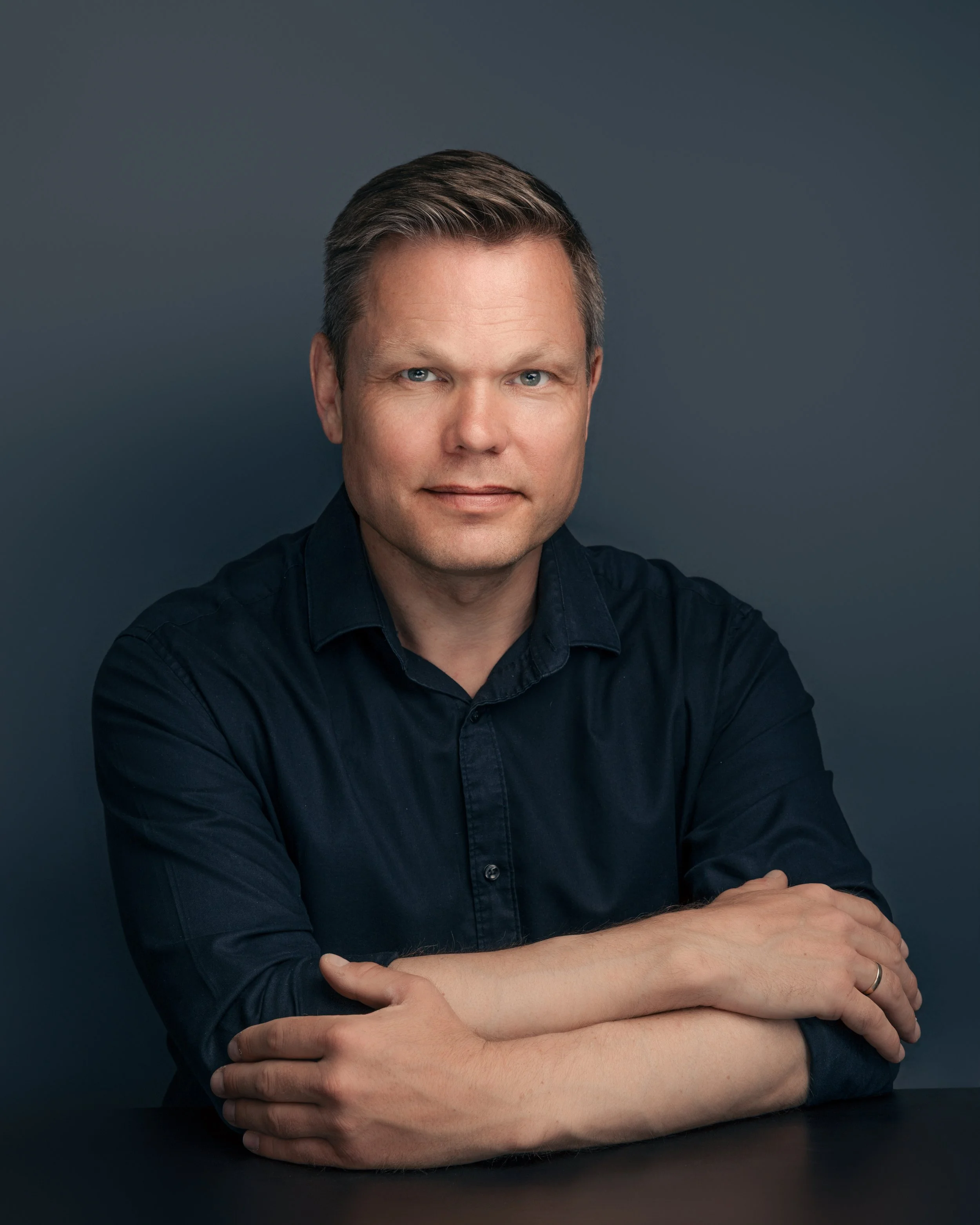 Portrett av en mann med kort hår, iført en mørk blå skjorte, sittende med armene foldet foran en mørk blå bakgrunn.