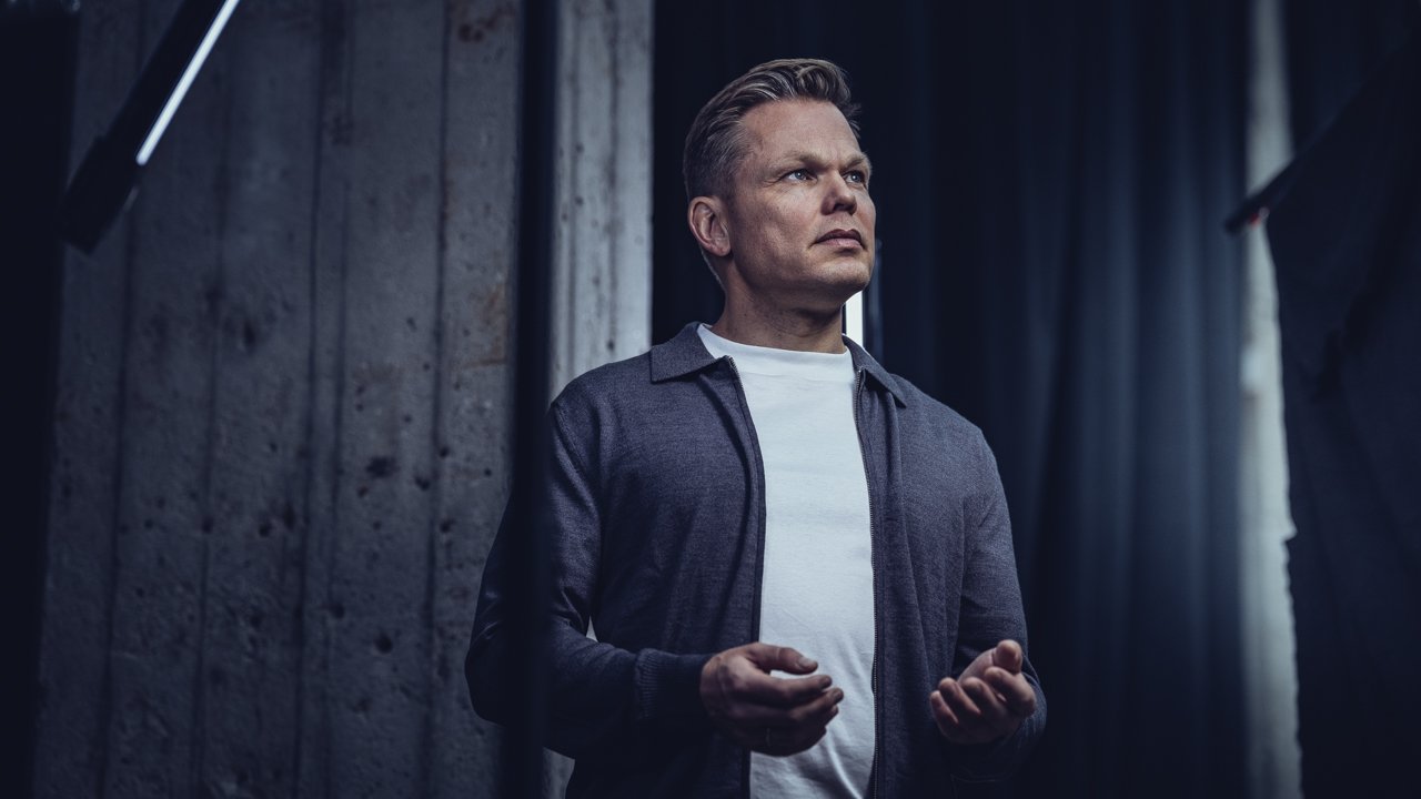 En mann med blond hår, iført en grå jakke og hvit T-skjorte, står i et mørkt rom med en betongvegg og profesjonell belysning.