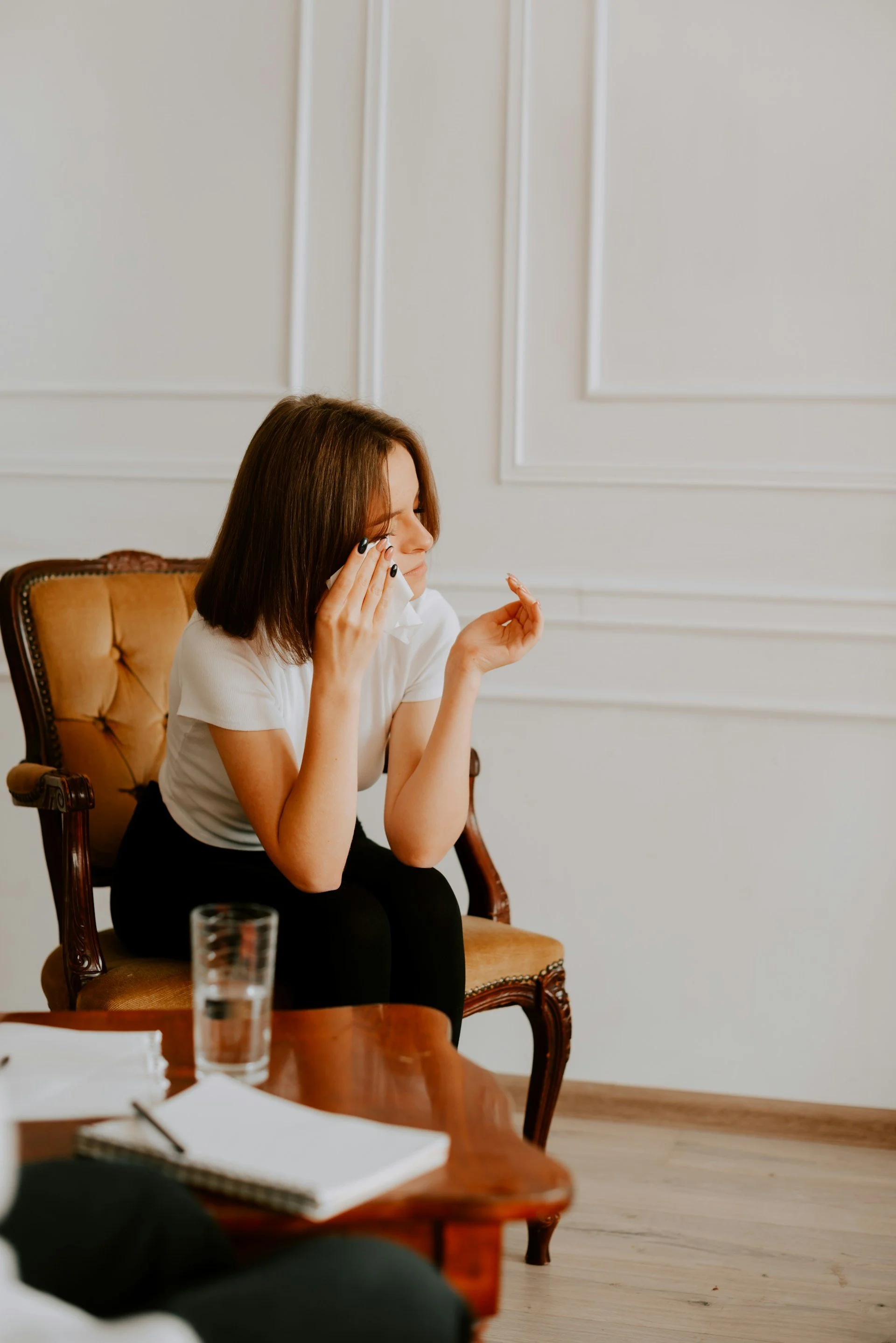 A woman sitting on a wooden chair with a tan upholstered back, wiping her eye with a tissue, wearing a white shirt and black pants, in a room with white paneled walls, a glass of water, and a notebook on a wooden table.