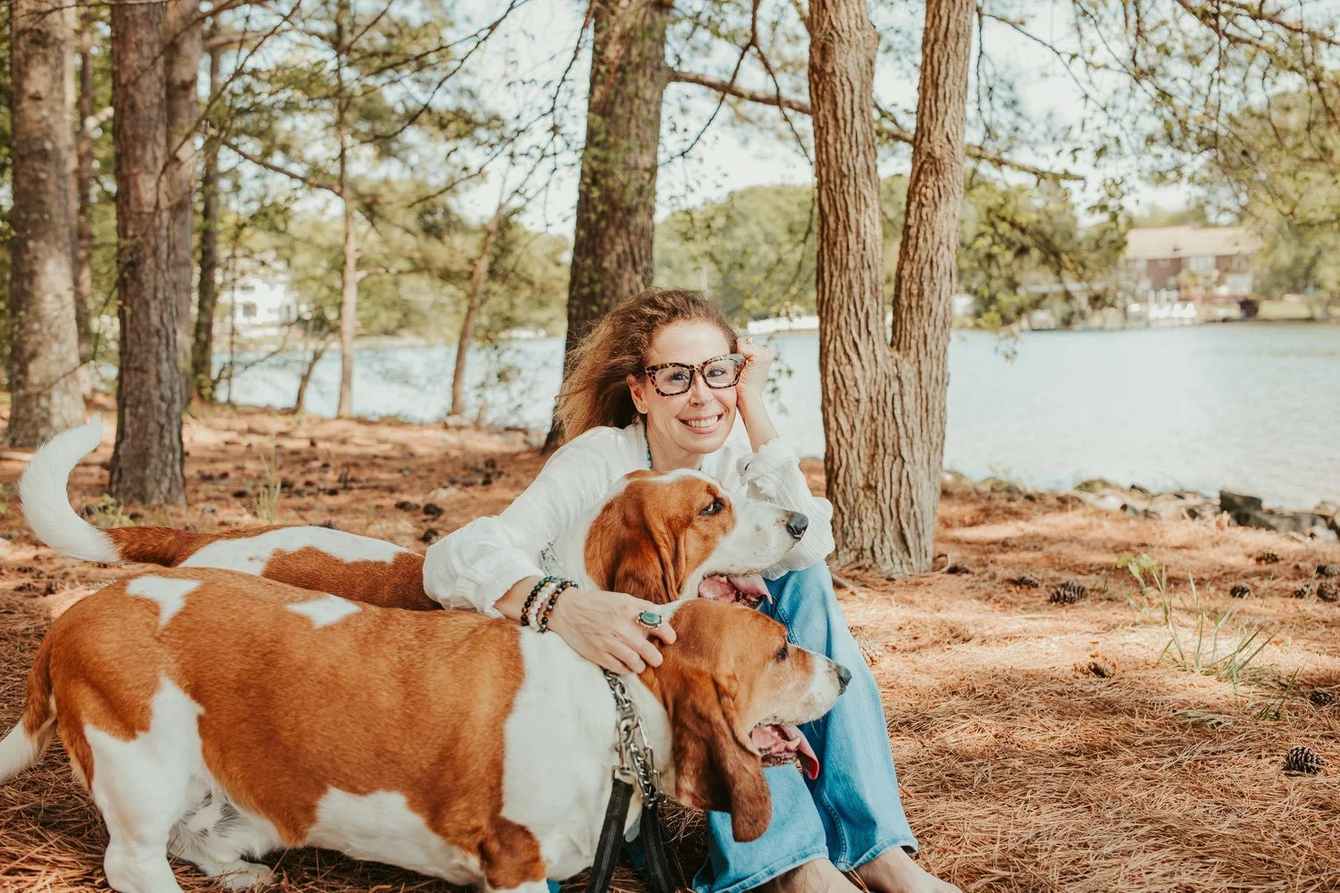 Woman sitting on forest ground by lake with three basset hound dogs, trees in background, smiling at camera.