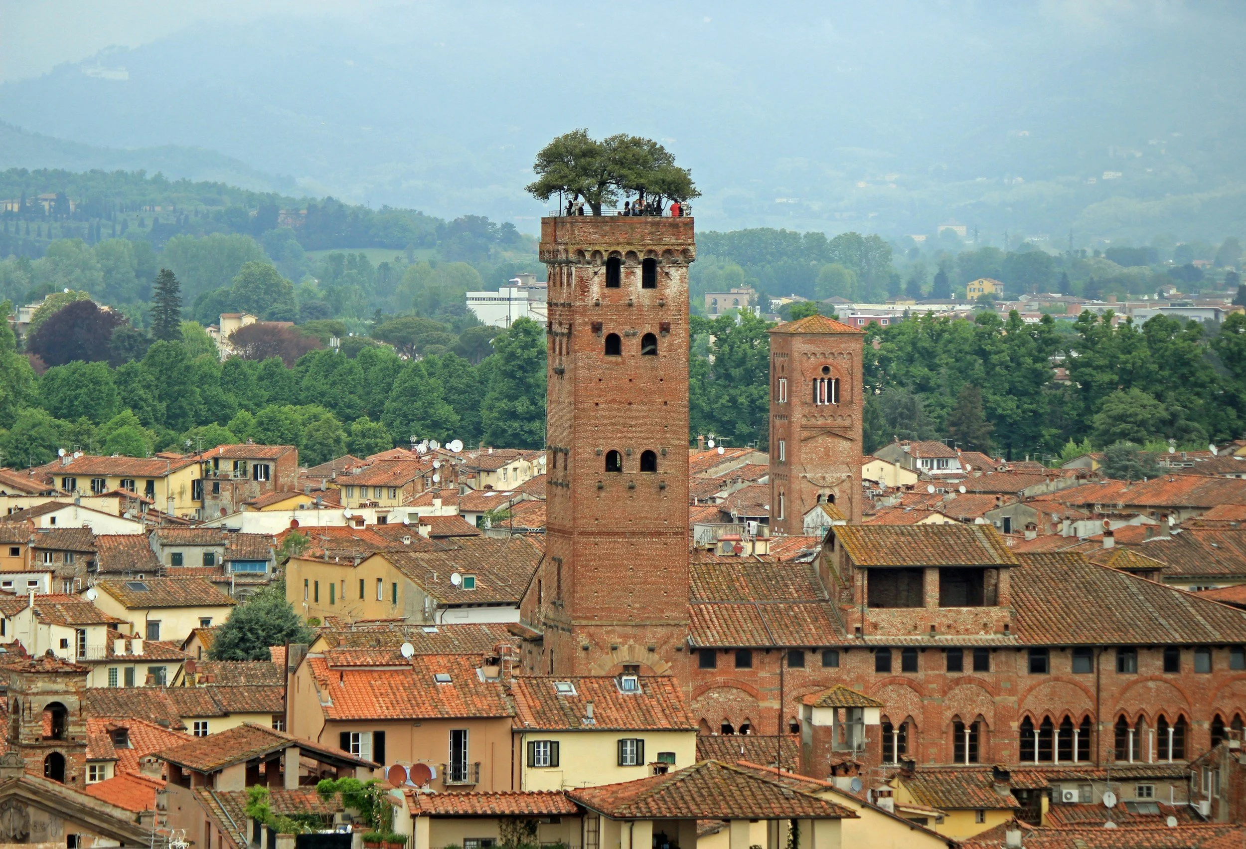 Aerial view of Lucca, Italy, with the Torre Guinigi tower and its rooftop oak garden rising above the Street decorated with Union Jack flags and red, white, blue pennant banners, with shops and people walking, and the ocean visible in the background.