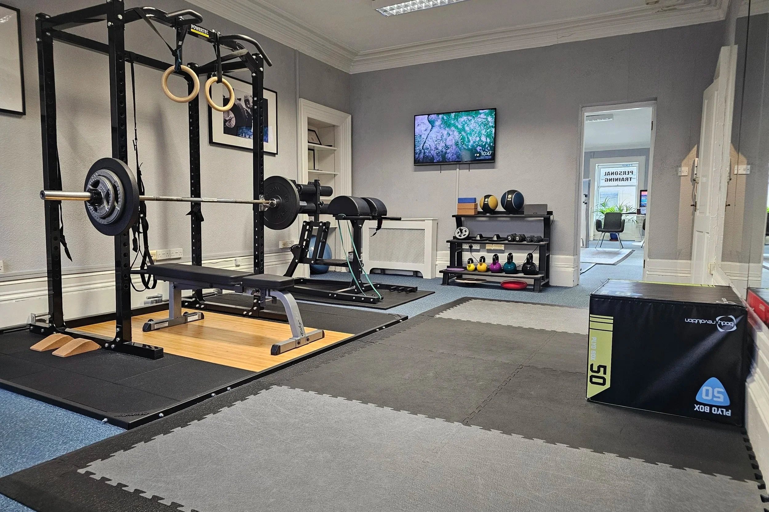 Home gym with black power rack, bench, dumbbells, kettlebells, gym mats, and a TV on a gray wall.