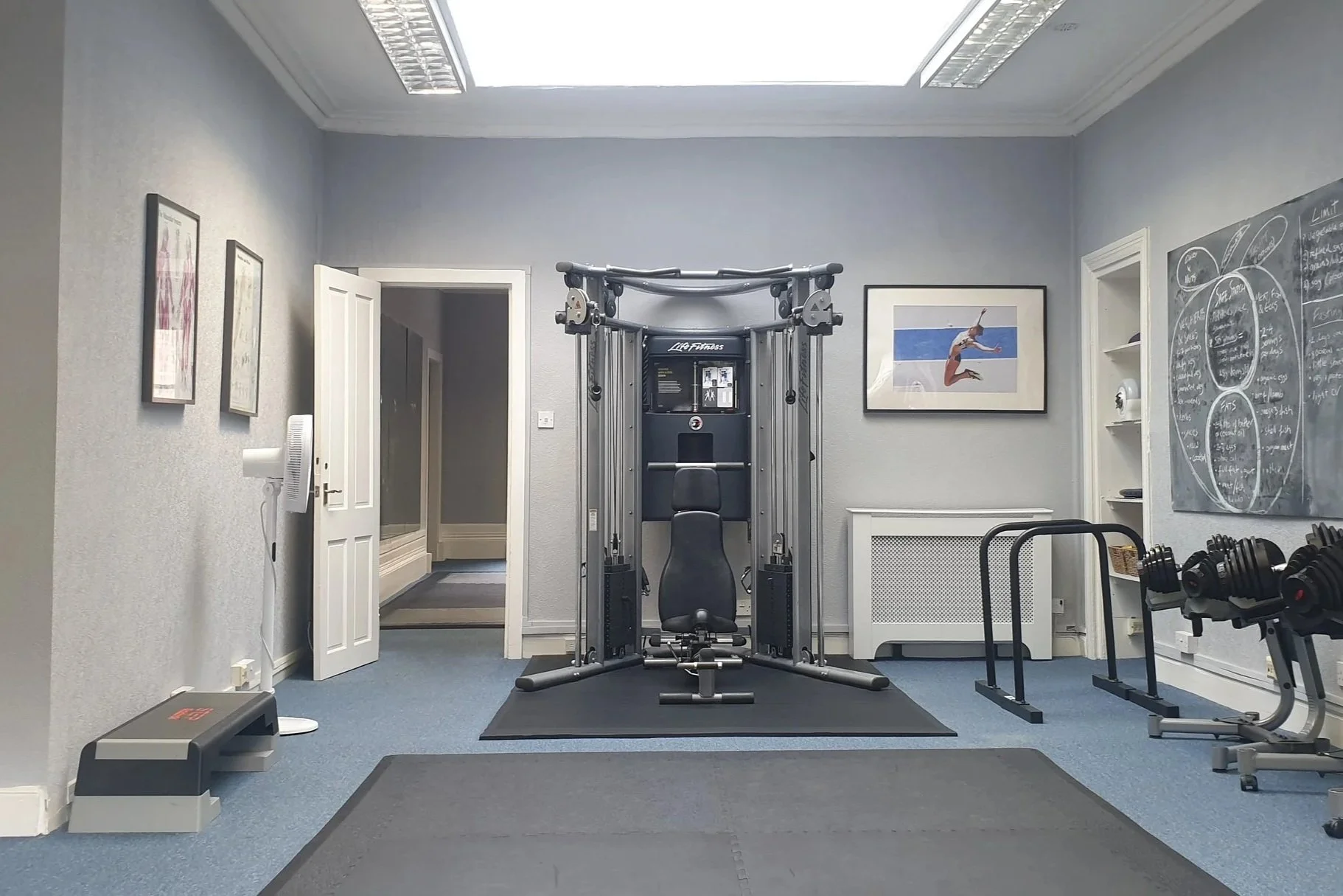 A home gym room with exercise equipment including a multi-functional workout station, two sets of dumbbells on a rack, a step platform, a black textured floor mat, and wall decor including framed pictures and a large blackboard with handwritten notes