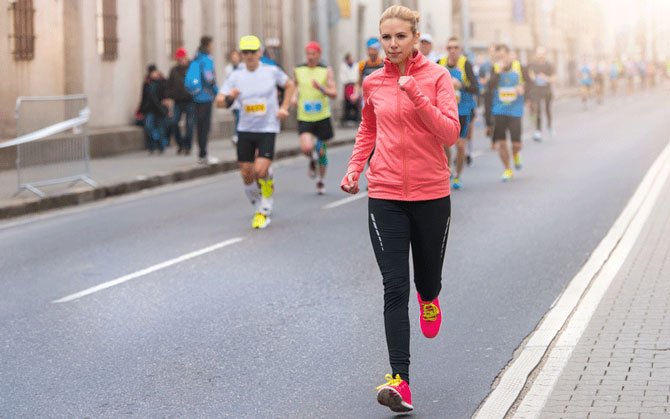 A woman in a pink jacket and black athletic pants running on a city street during a marathon or race, with other runners and spectators in the background.