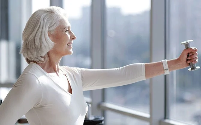 An elderly woman with gray hair exercising with a dumbbell in front of a large window.