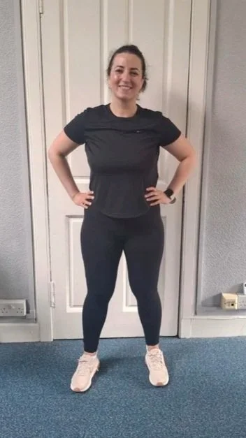 A woman standing with hands on her hips, smiling, wearing a black t-shirt, black leggings, and white sneakers in front of a closed door.