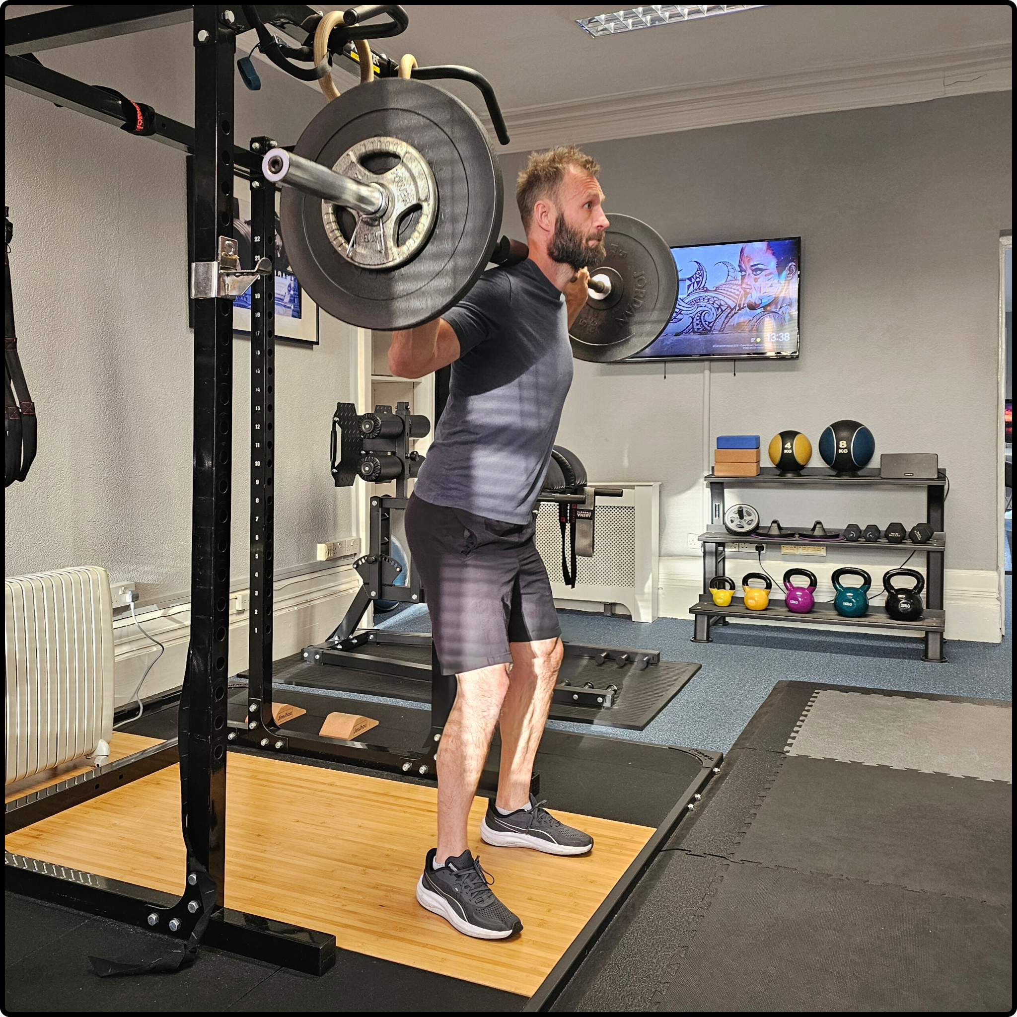 A man lying on a workout bench, lifting dumbbells in a gym, with a chalkboard and workout notes in the background.