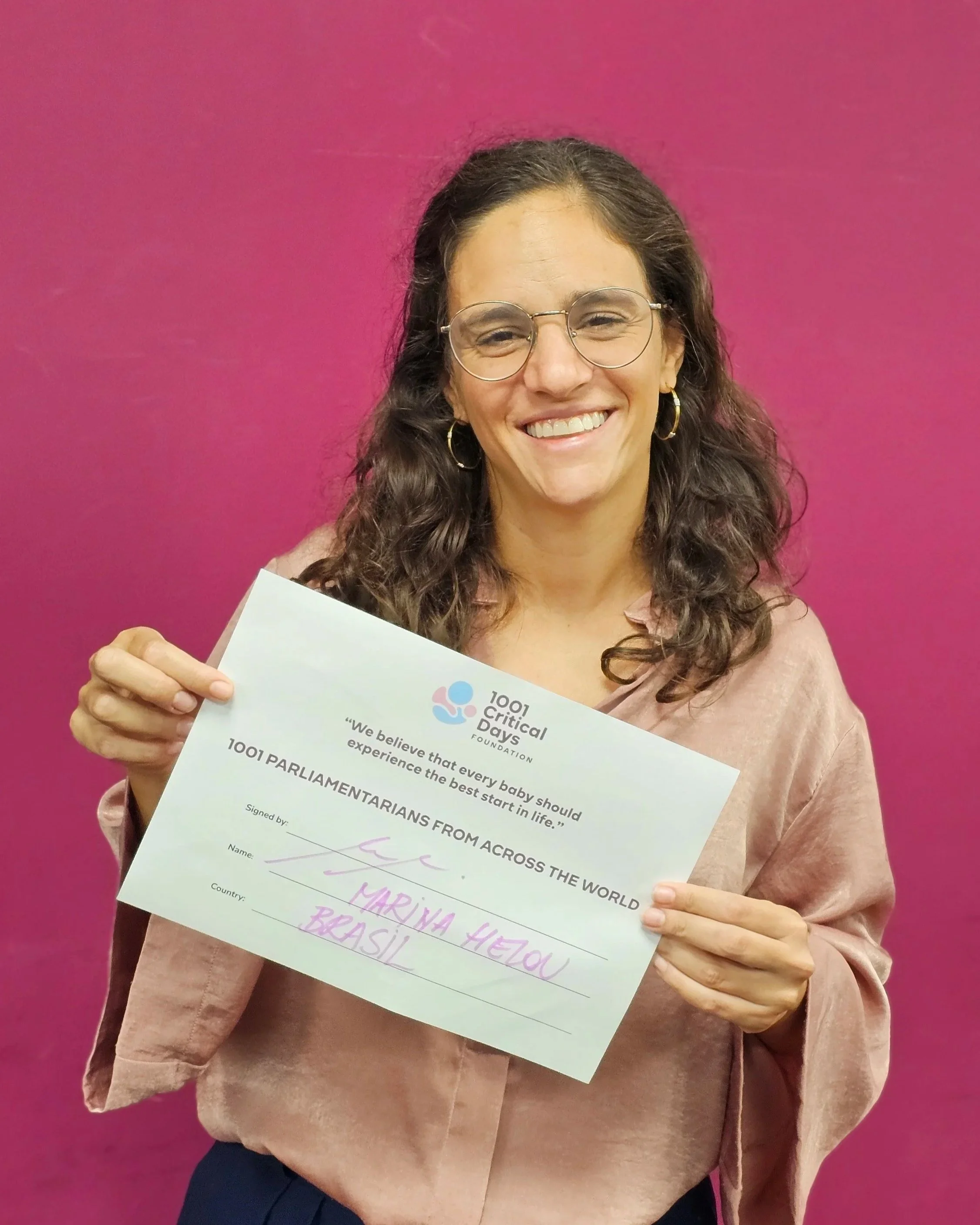 A woman smiling and holding a sign related to 1001 Critical Days Foundation, standing in front of a pink wall.