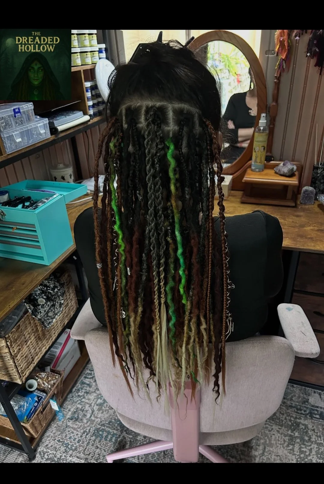 Back view of a person with long, colorful, and textured dreadlocks sitting in a salon chair in front of a mirror.