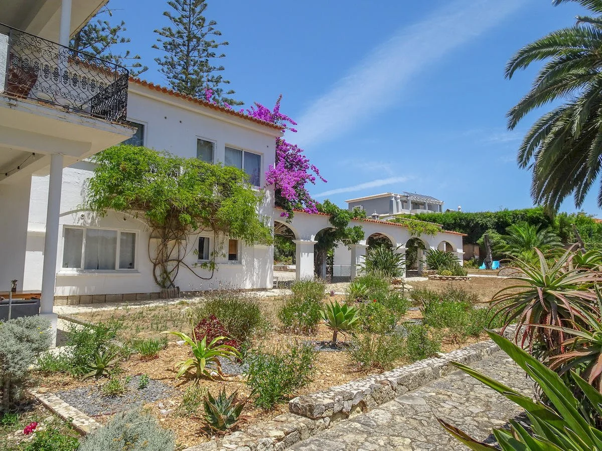 White Mediterranean-style house with arched windows and balconies, surrounded by lush greenery, flowering plants, and palm trees under a blue sky.
