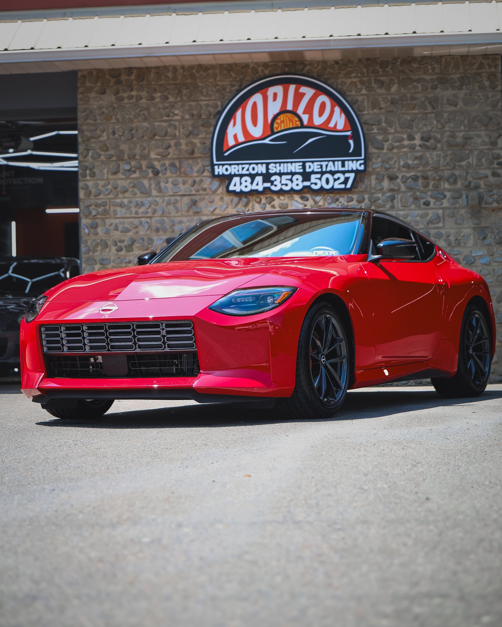 Red sports car parked in front of a brick building with a sign that reads 'Horizon Shine Detailing' and a phone number.