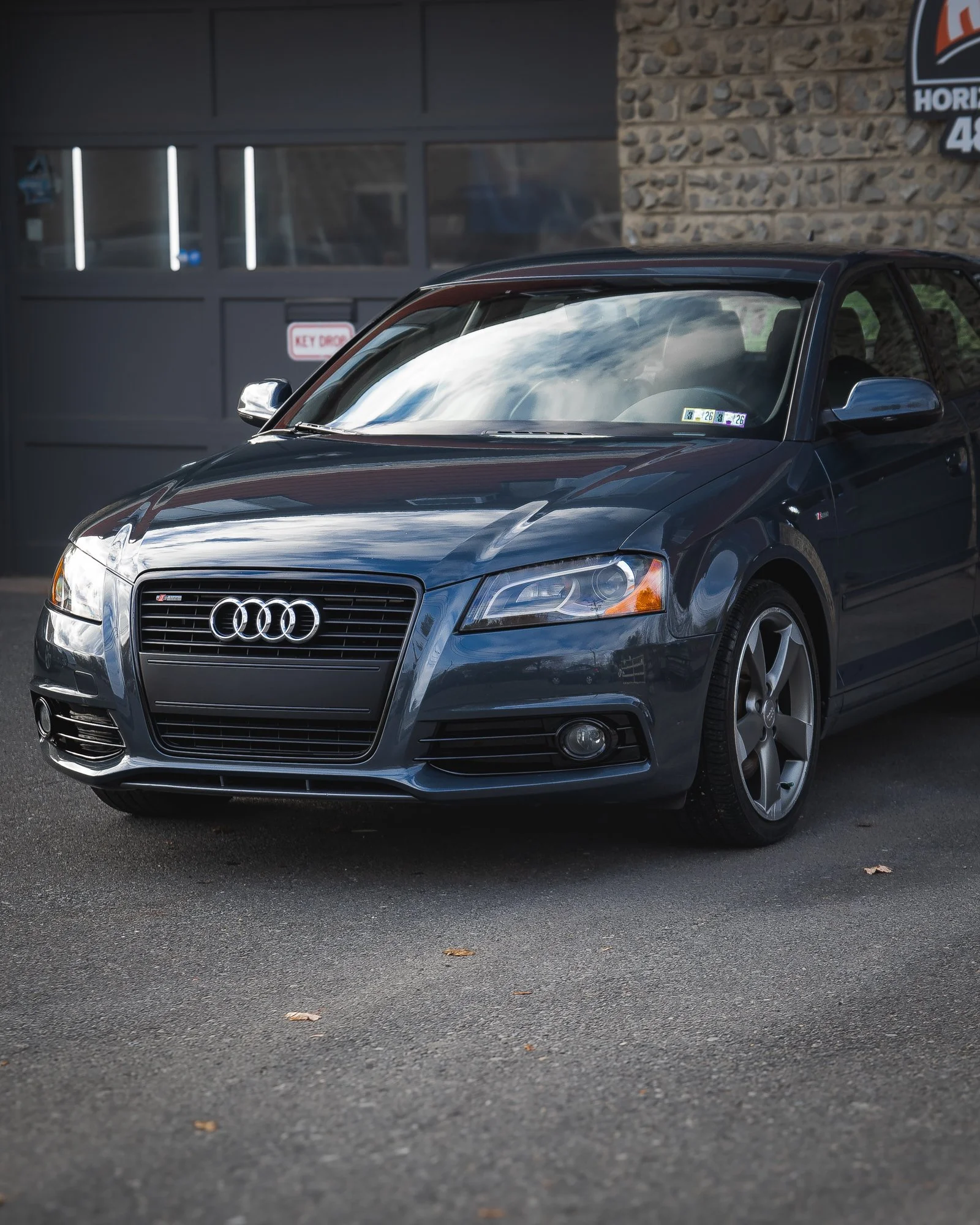 Black Audi sedan parked outside near a building with stone and metal siding, under a cloudy sky reflection on the windshield.