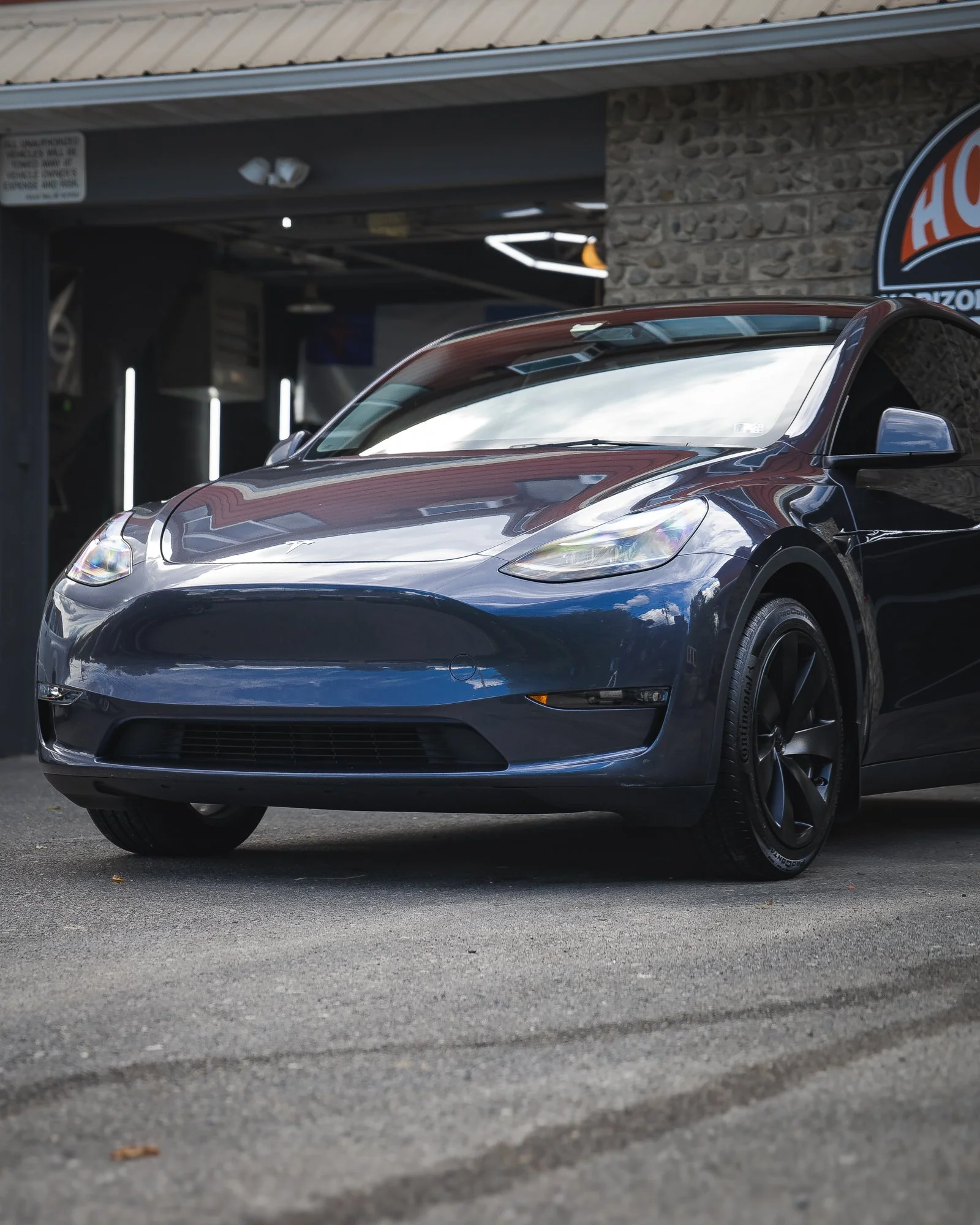Front view of a dark blue Tesla Model 3 parked outside a building with a stone wall and a partially visible propane sign.