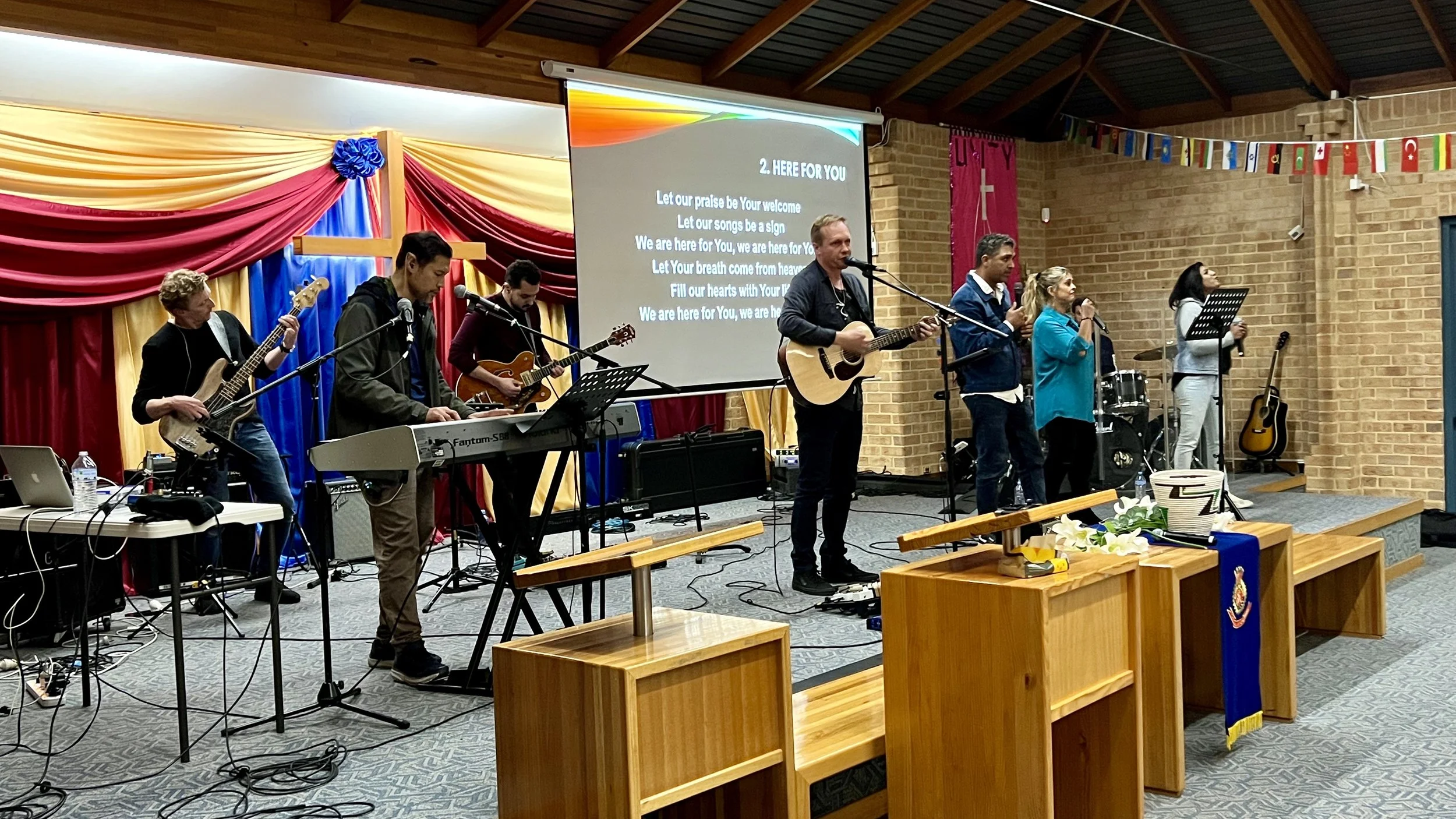 A worship band performing inside a church. Musicians are playing instruments such as a keyboard, guitars, and a bass. Vocalists are standing and singing near microphones. A projection screen displays song lyrics. Decor includes flags and fabric drapery against the walls.