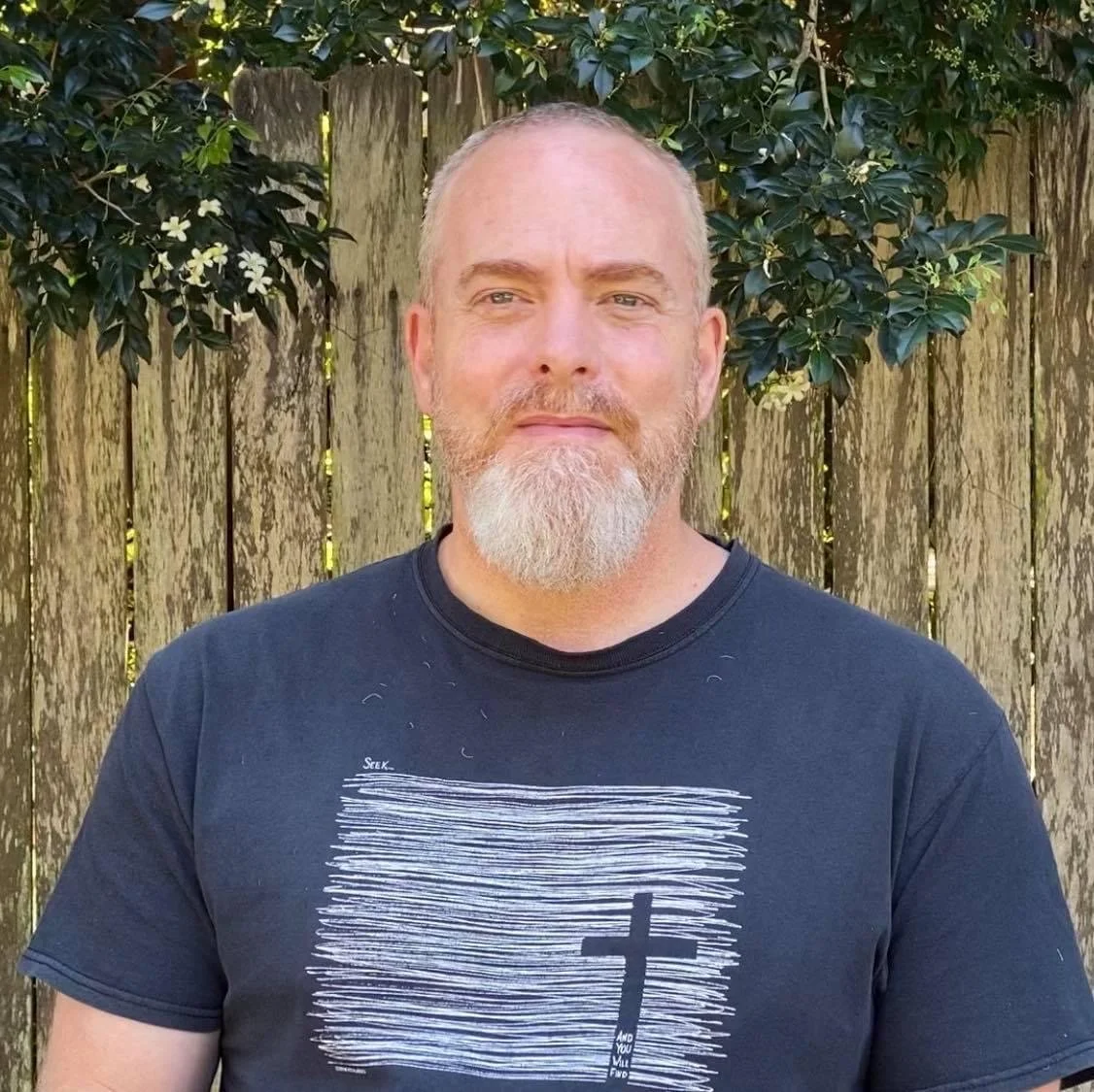 Man with a beard wearing a black T-shirt with a cross design stands in front of a wooden fence and greenery.