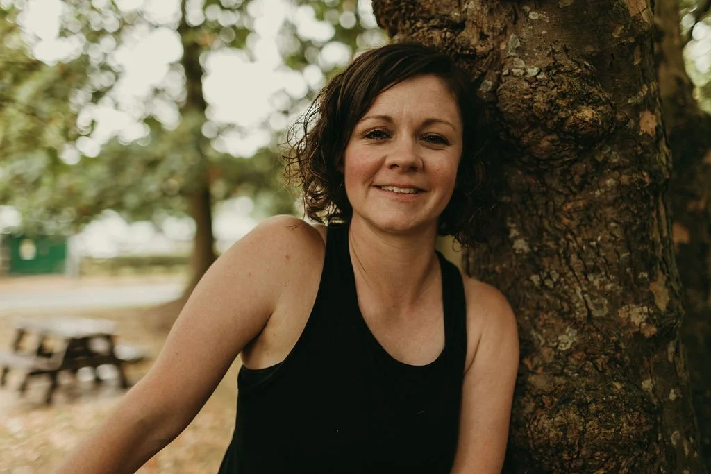 Young woman smiling outdoors wearing a straw hat, black crop top, and patterned skirt, with tall grass and trees in the background.