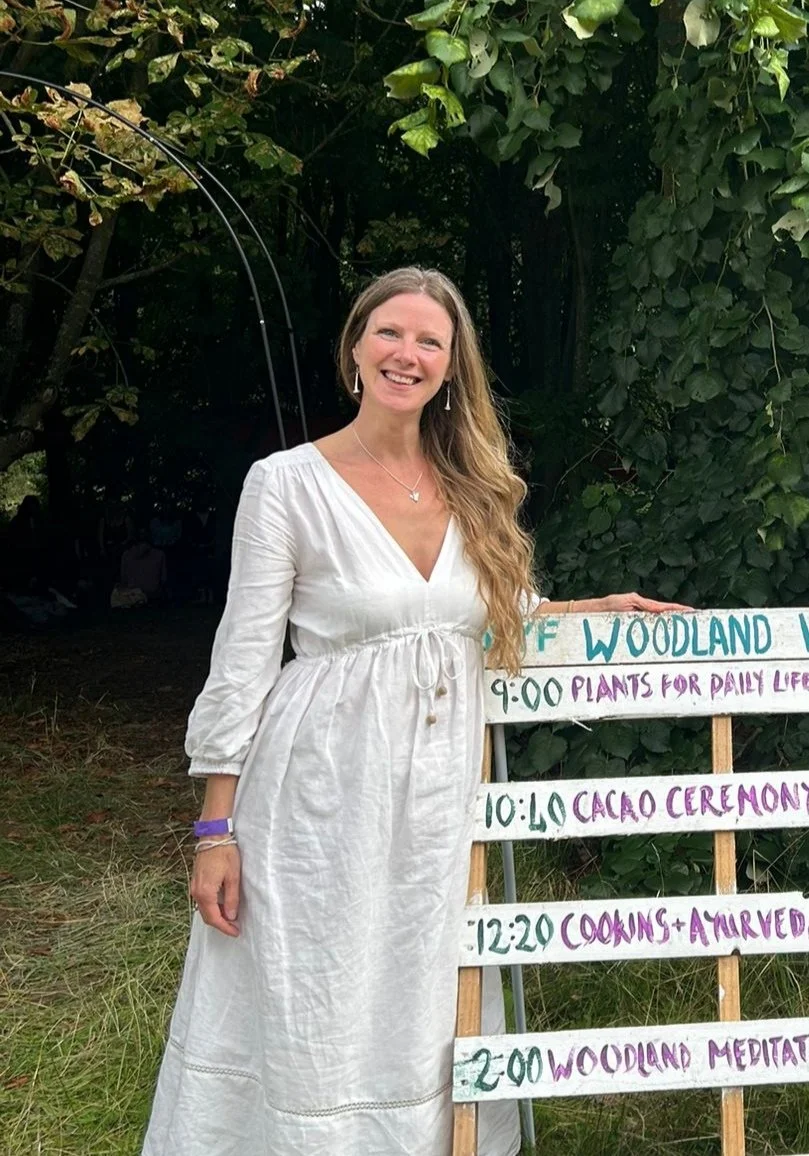 A woman in a beige dress standing on a wooden balcony surrounded by lush green tropical plants.