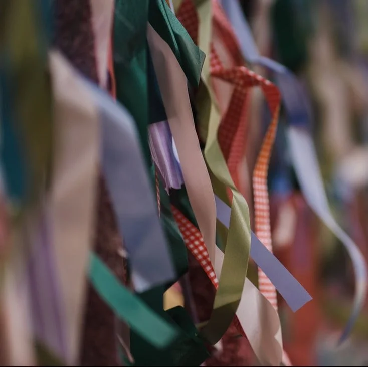 A woman in a yoga studio or meditation space arranging flowers in a vase, surrounded by candles, small statues, and decorative plants, with a large chakra symbol on the wall behind her and sunlight streaming in.