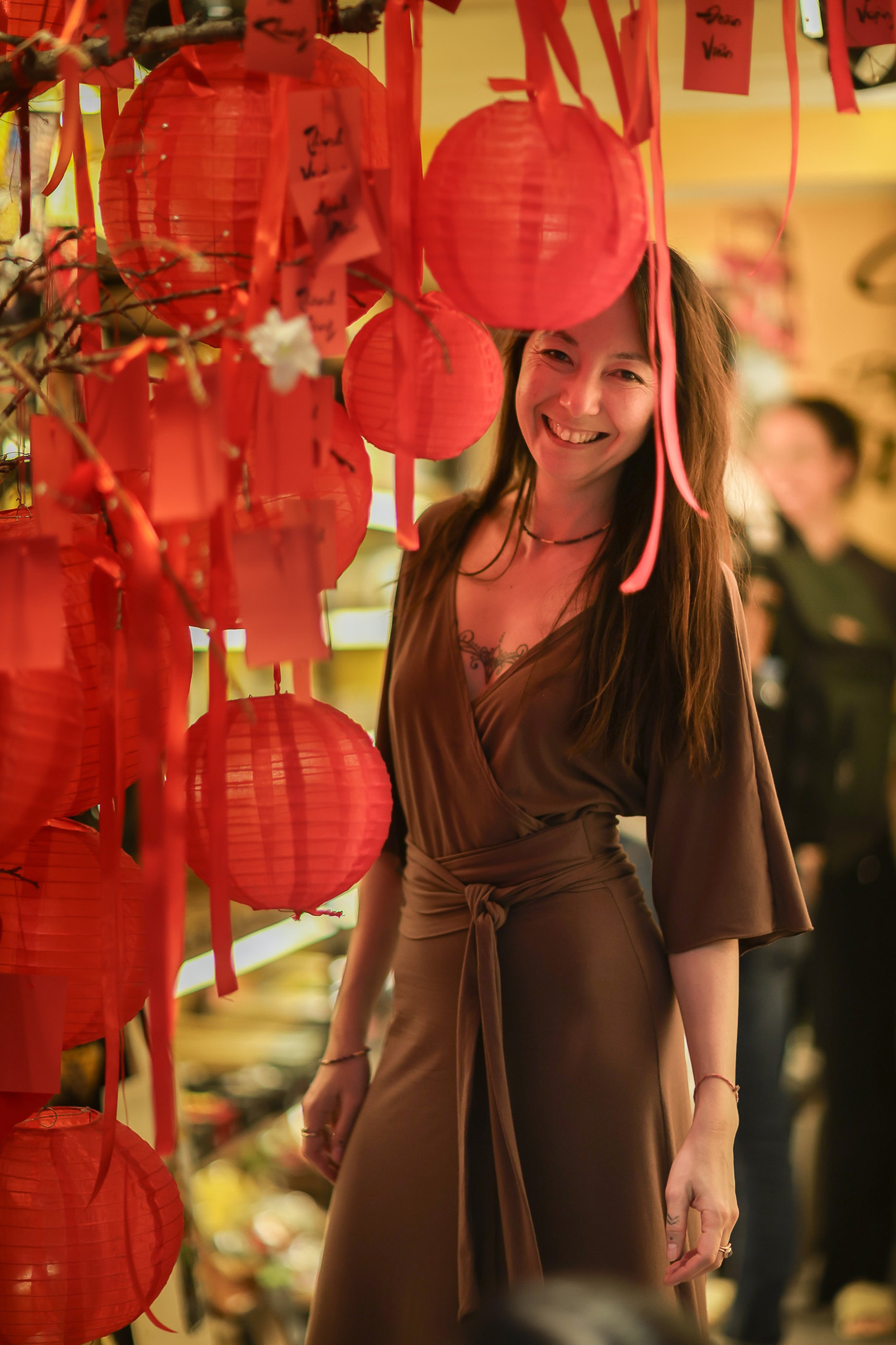 A woman with curly brown hair smiling, wearing a pink, purple, and light blue windbreaker, indoors in a well-lit room with a bookshelf, framed photos, and a white lamp in the background.