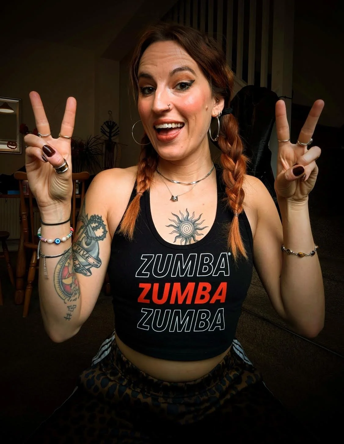 A woman with red hair wearing a purple t-shirt and black pants, flexing her right arm and smiling, against a black background.
