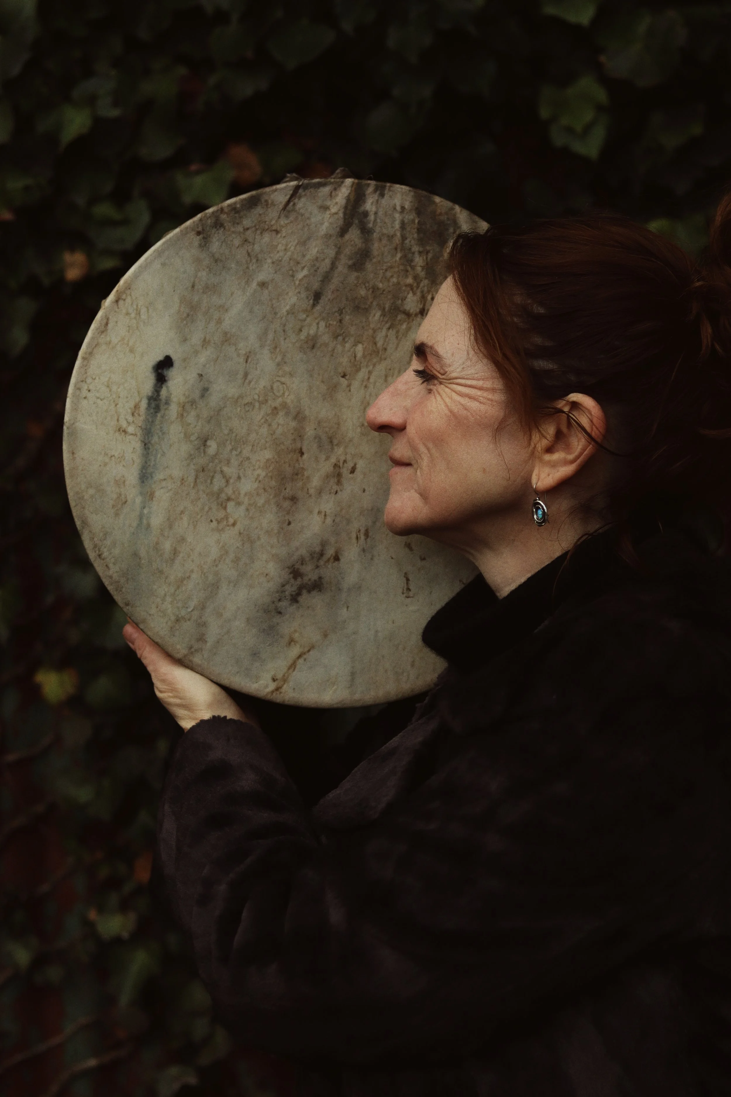 A woman in a white, eyelet dress sits cross-legged on a blue and white blanket outdoors, surrounded by green foliage, playing a crystal singing bowl with other similar bowls nearby.