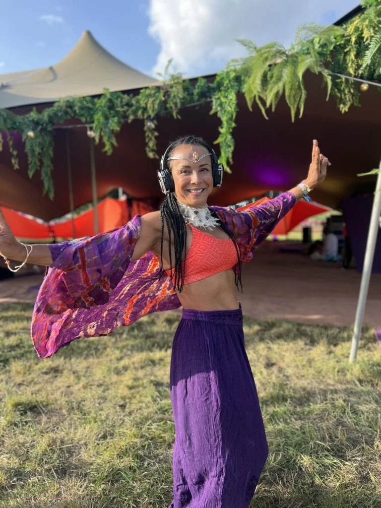 A smiling young woman with glasses on her head and a nose ring, standing outdoors with trees and a dirt ground in the background, wearing a black shirt and holding a strap of her bag.