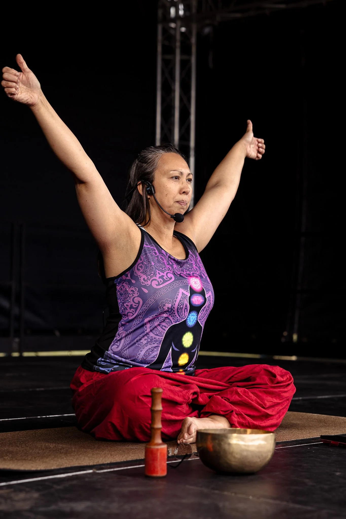 Woman sitting cross-legged on a yoga mat, smiling, with singing bowl and mallet nearby, in a well-lit room with large windows and radiators.