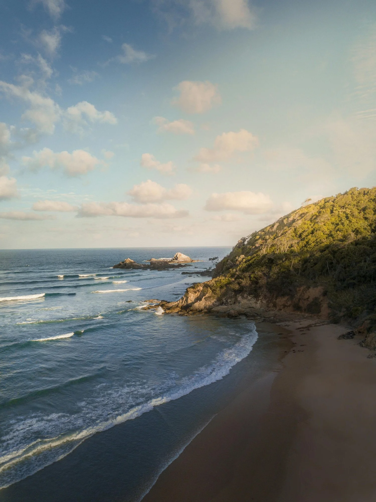 Beach in Byron Bay with waves crashing on the shore, rocky formations in the water, and a hillside covered with trees and vegetation under a partly cloudy sky.
