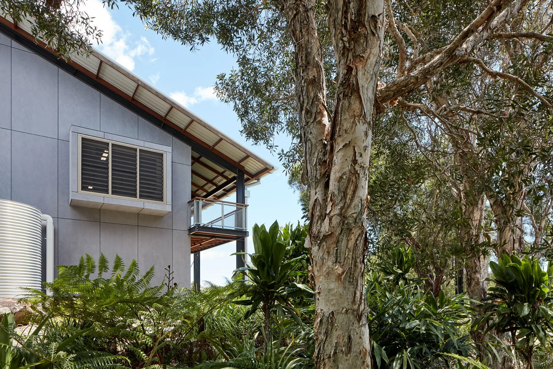 View of a modern commercial building with gray exterior walls, a balcony with a glass railing, and a sloped roof. There are large trees and lush green plants in the foreground under a partly cloudy blue sky at Habitat Byron Bay.