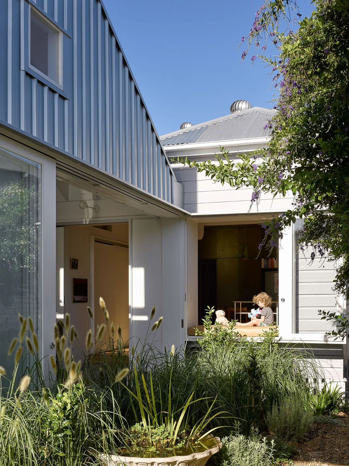 A young child sitting in a sunlit outdoor room, playing with plush toys, with a lush garden and plants in the foreground, and a modern building with a metal roof in the background.