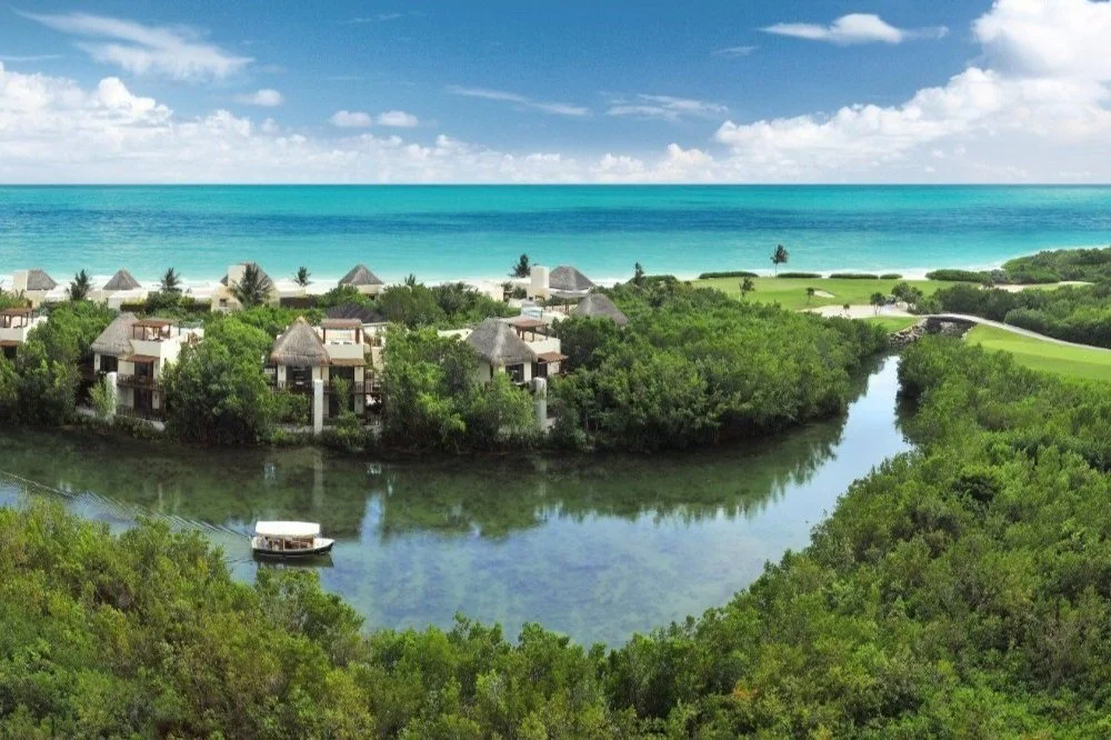 Tropical resort with thatched-roof villas, lush greenery, calm river, and ocean in the background under a partly cloudy sky.