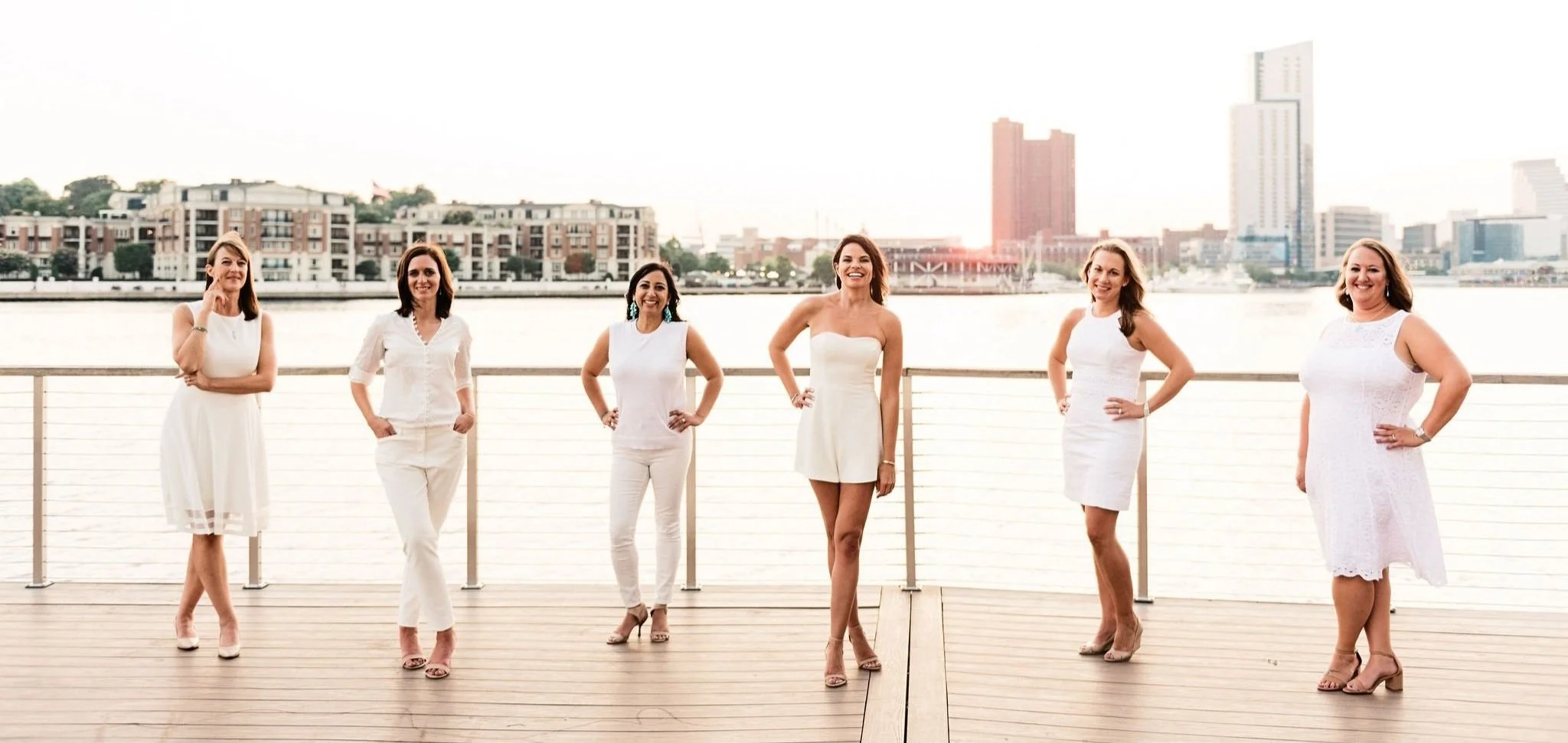 Six women dressed in white standing on a wooden deck by a city waterfront at sunset.