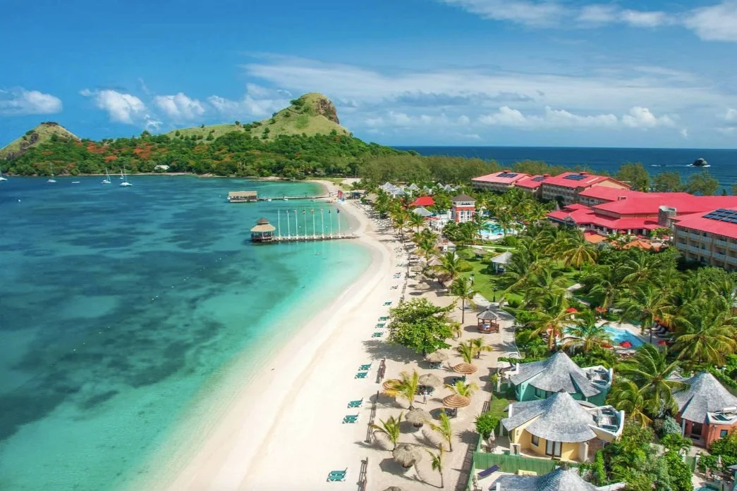Aerial view of a tropical beach resort with white sands, turquoise water, palm trees, and multiple buildings with red roofs, set against a green hill and ocean in the background.