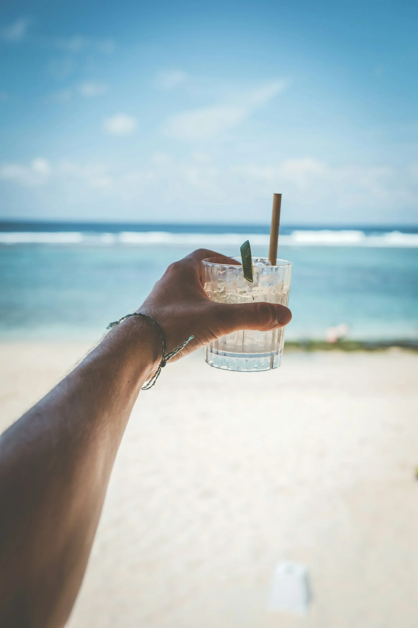 A person holding a clear glass with a beverage and lime wedge, overlooking a beach and ocean with blue sky and scattered clouds.